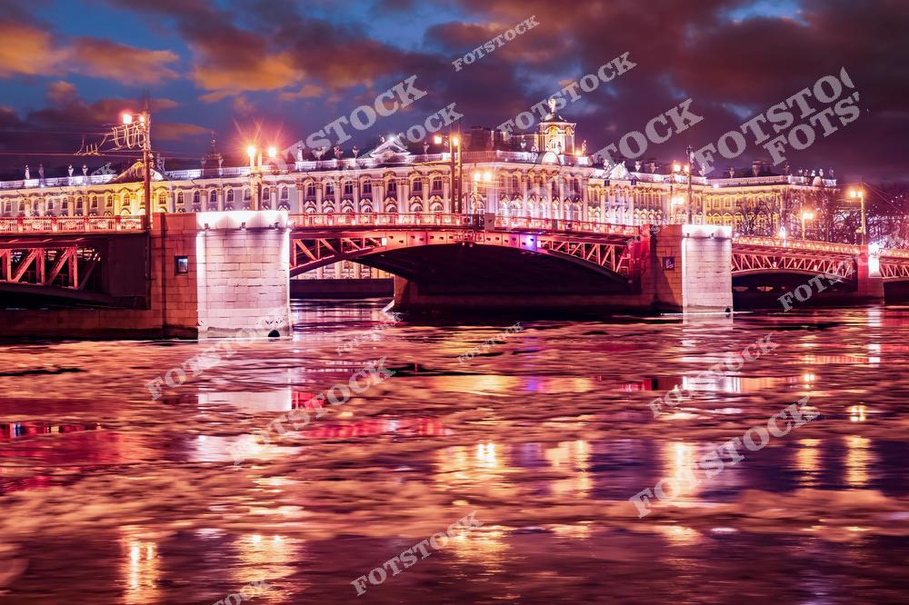 На снимке изображен Дворцовый мост в Санкт-Петербурге, освещённый яркими огнями в вечернее время. Водная гладь реки Невы покрыта плавающим льдом, что добавляет ощущение зимнего холода. Архитектурная красота моста и окружающих зданий подчёркивается отражениями в воде. Это место — одна из визитных карточек города.